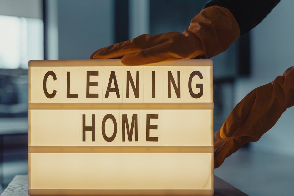 A person wearing brown gloves is adjusting a lightbox display with the words 'CLEANING HOME' printed on it, indoors with soft lighting. The background shows a modern interior with a blurred window or