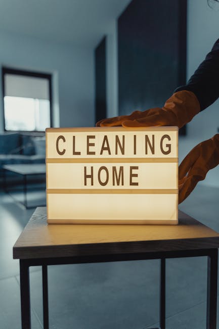 A person wearing brown gloves is adjusting a lightbox display with the words 'CLEANING HOME' printed on it, indoors with soft lighting. The background shows a modern interior with a blurred window or